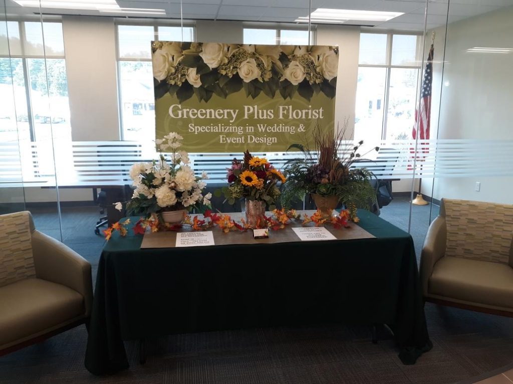 Business display table for Greenery Plus Florist decorated with flowers and signs.